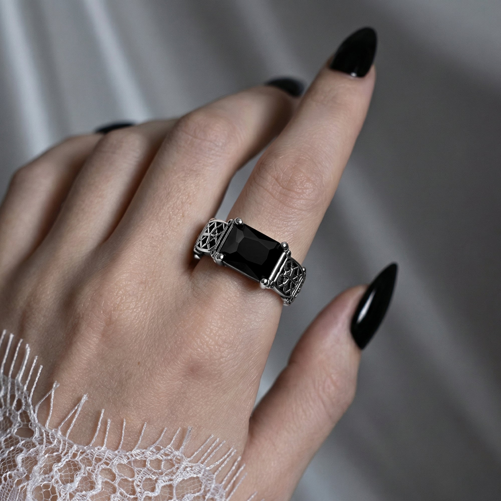 Obsidian gothic cross ring with black stone worn on hand with black nails and white lace sleeve against grey background